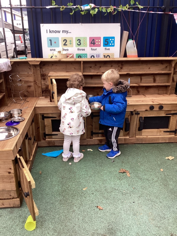 Two children playing with a wooden microwave in the outdoor kitchen