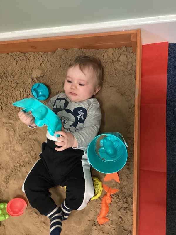 A toddler lies in a sand pit holding and surrounded by toys