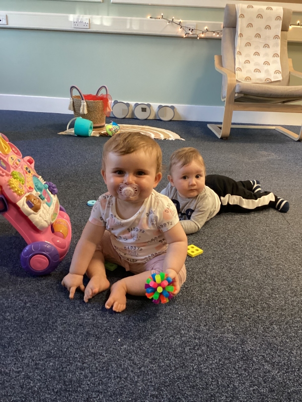 Two toddlers sit and lie on the floor with toys, looking at the camera